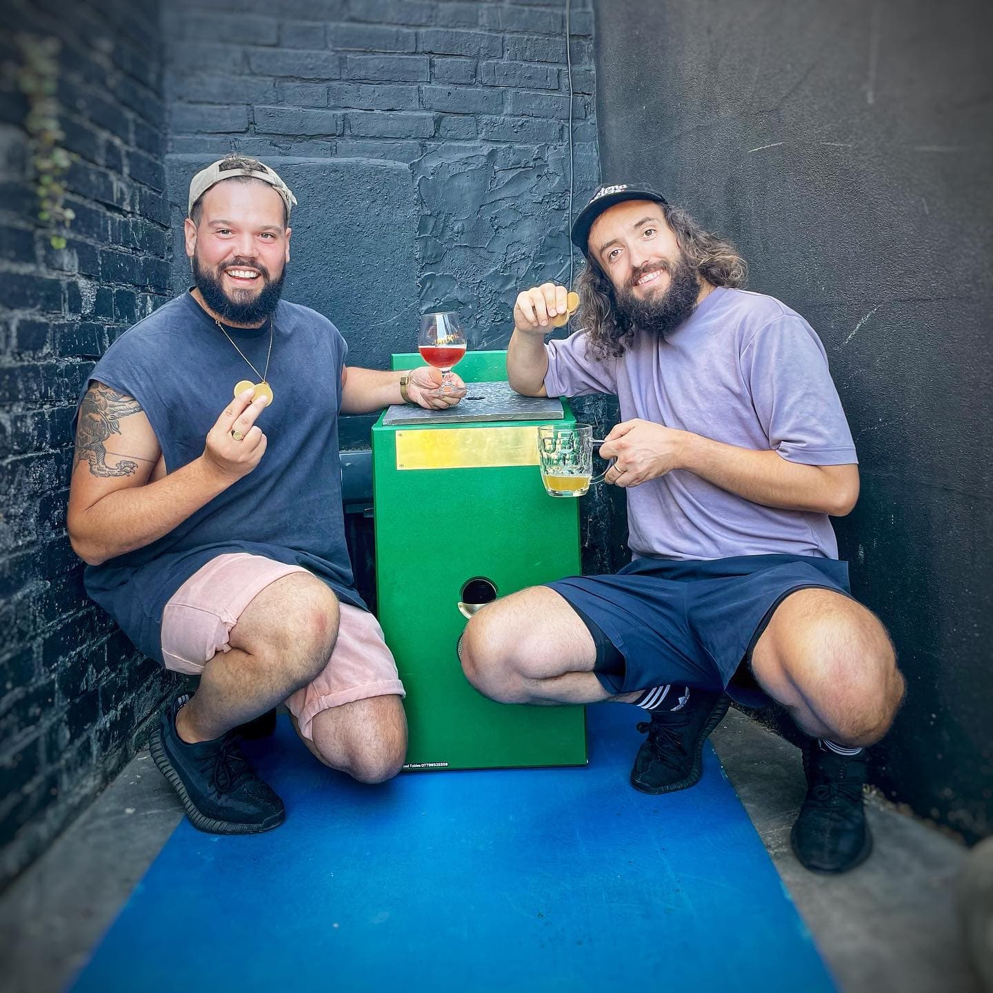 Alex and Sam beside their green Toads table, holding brass discs and drinks.